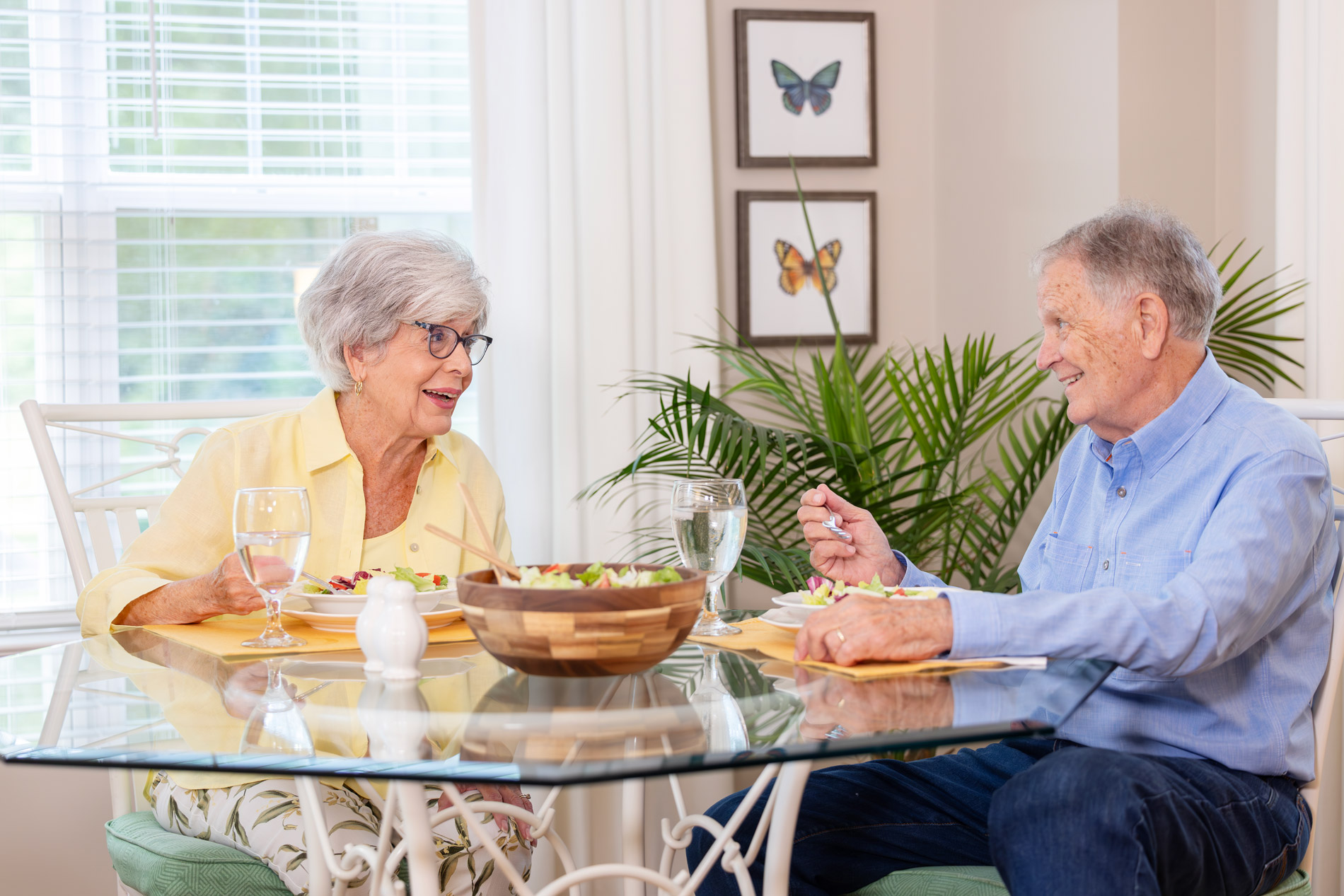 An elderly couple sits at a dining table, enjoying a meal together and smiling at each other.