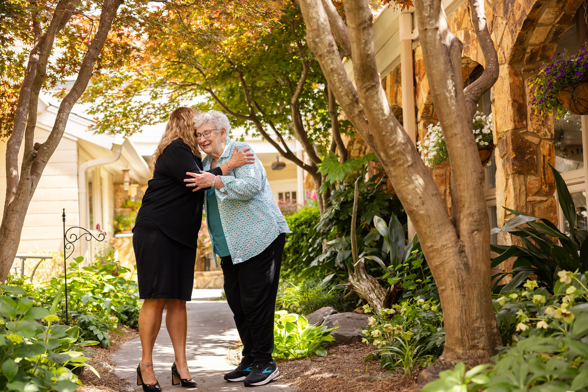 Two women happily hugging on a garden pathway, surrounded by trees and plants.