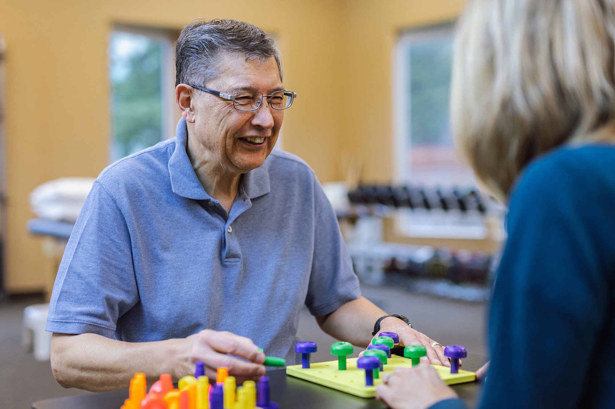 Man engaged in therapy activity involving colored pegs with a therapist at a table indoors.