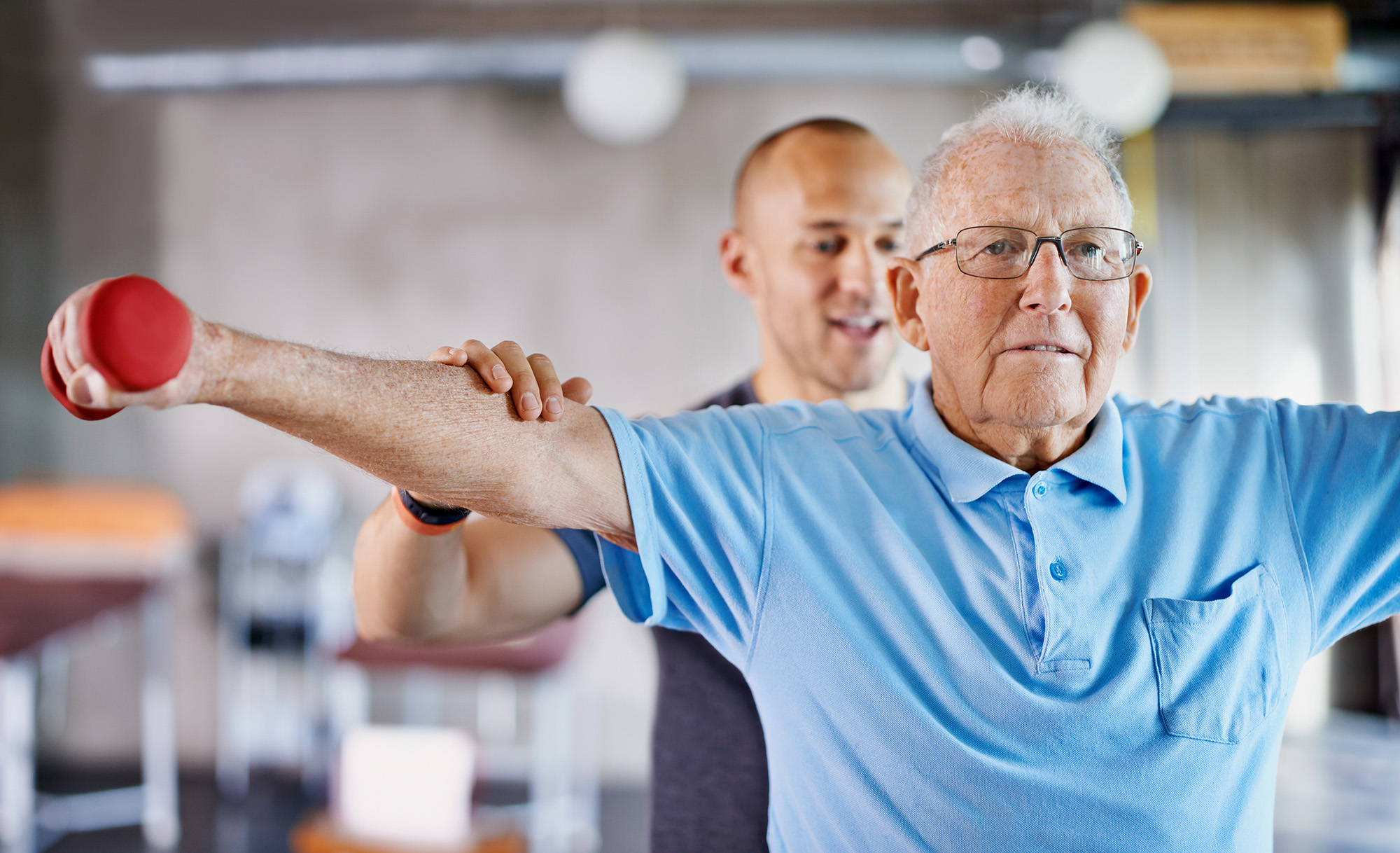 An older man lifts a red dumbbell while a trainer assists him in an exercise session.