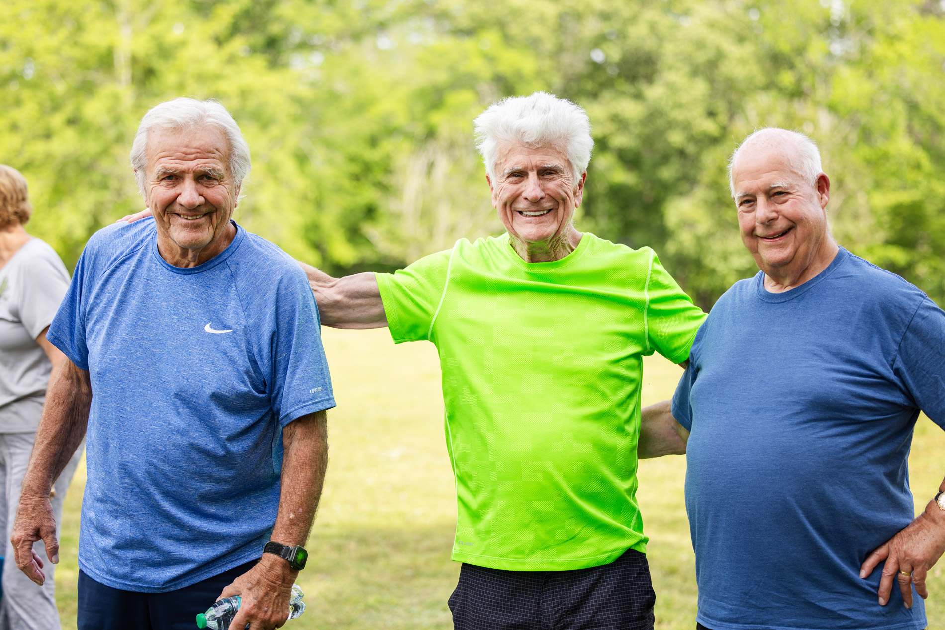 Three elderly men smiling, with their arms around each other, standing outdoors in casual clothing.