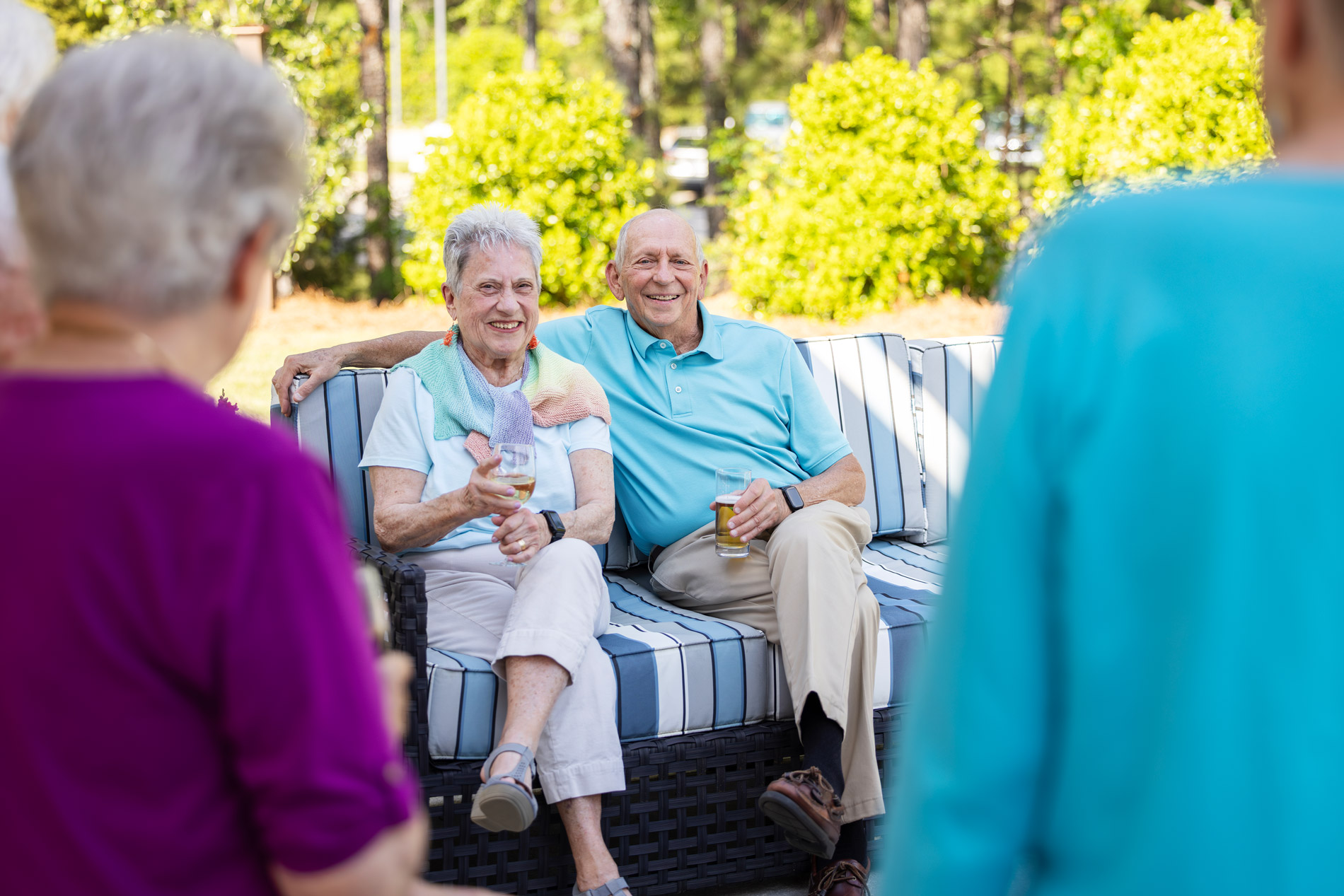 An elderly couple sitting on an outdoor sofa smiles while holding drinks at a social gathering.