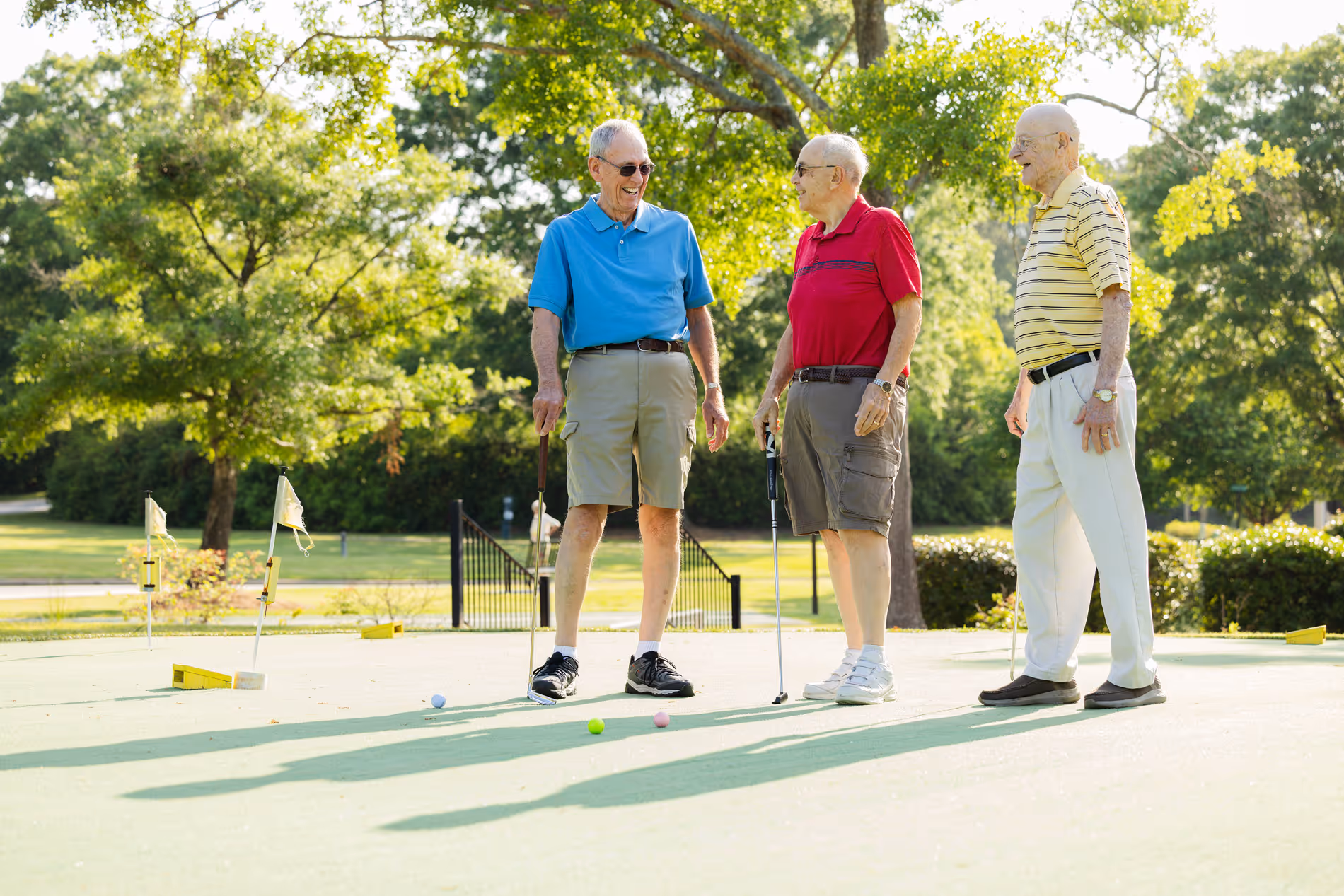 Three elderly men stand on a mini-golf course, talking and holding putters.