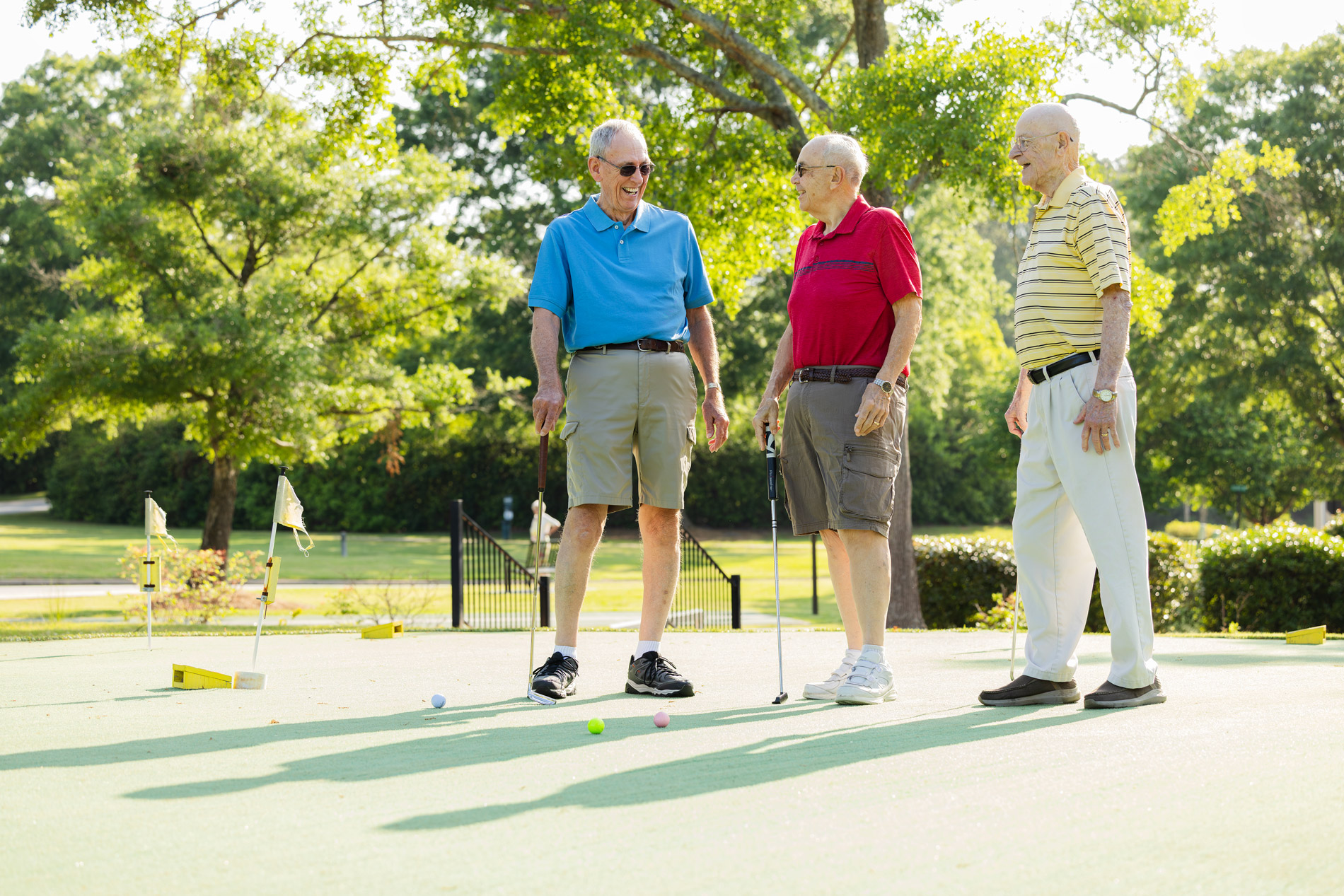 Three elderly men stand on a mini-golf course, talking and holding putters.
