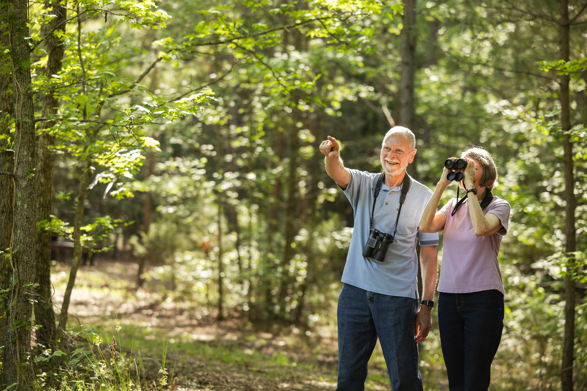 An elderly man points while a woman uses binoculars in a wooded area.