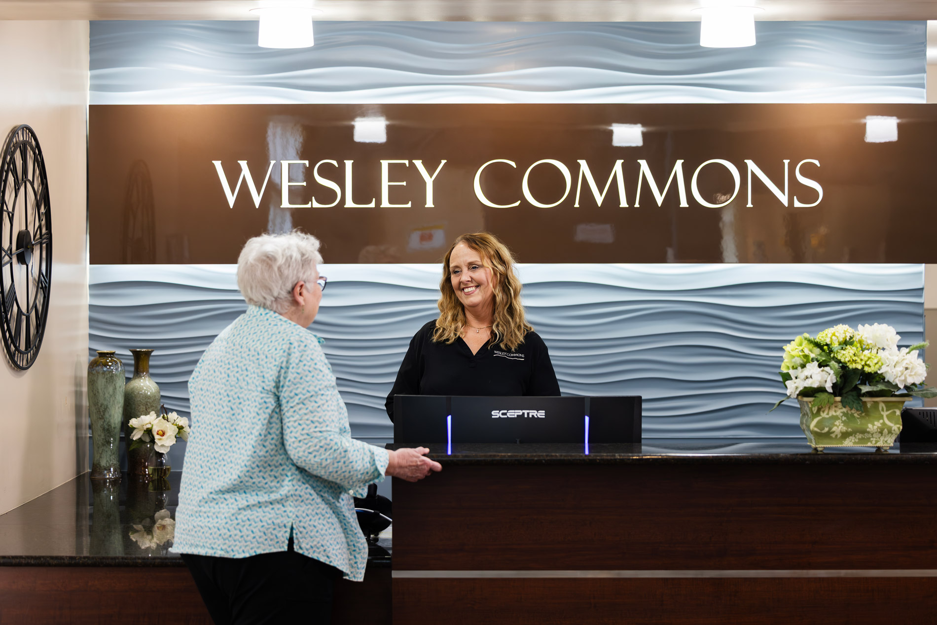 A staff member smiles while assisting an older woman at the Wesley Commons reception desk.