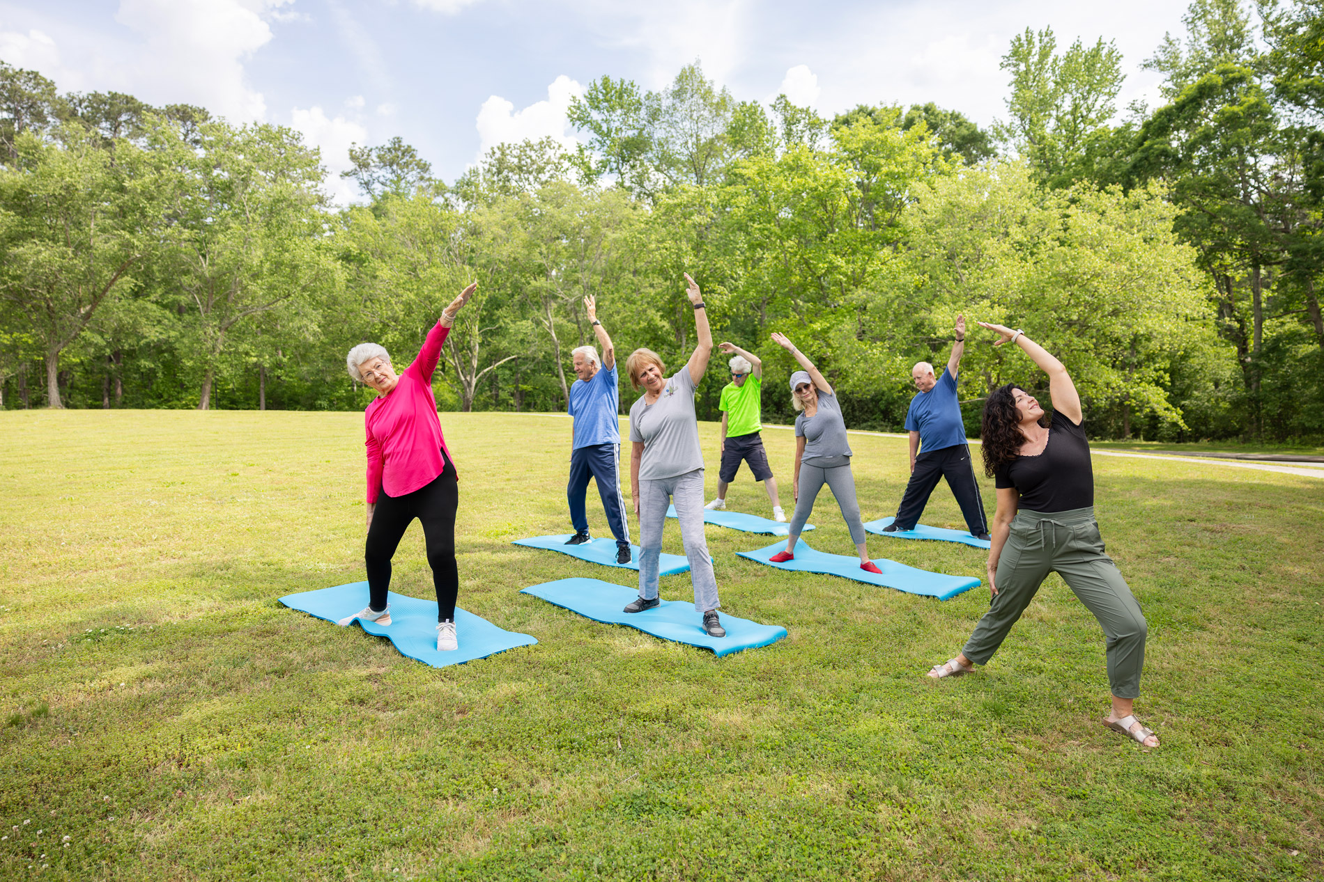 Seven people stretching on yoga mats in a park, raising one arm overhead for balance.