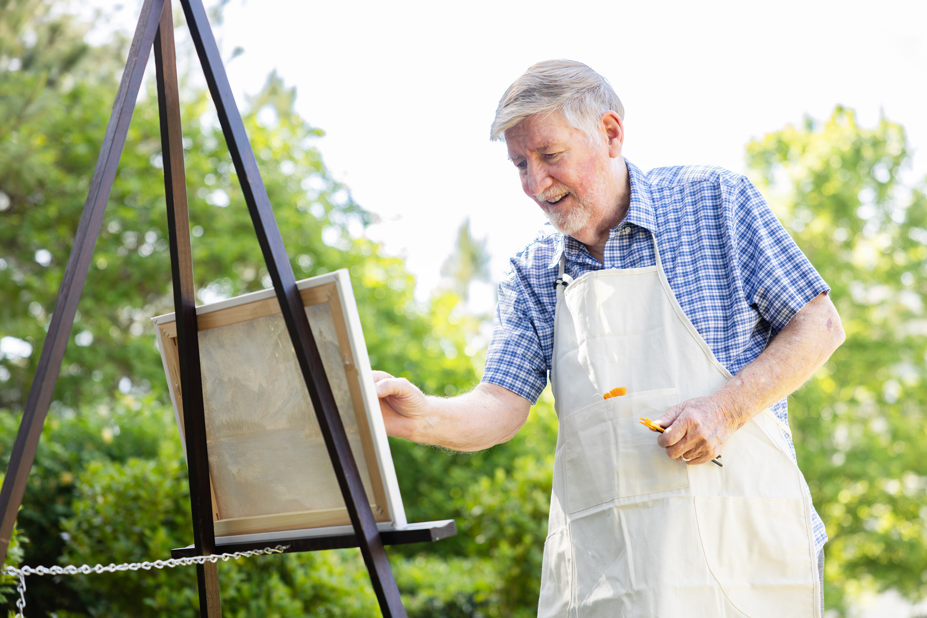 Elderly man painting on a canvas outside, wearing a checkered shirt and apron.