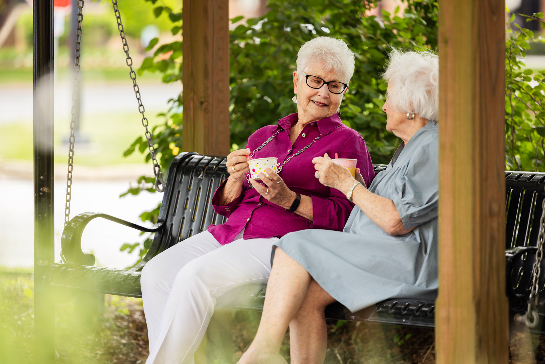 Two elderly women sit on a bench outdoors, enjoying a conversation with tea cups in hand.