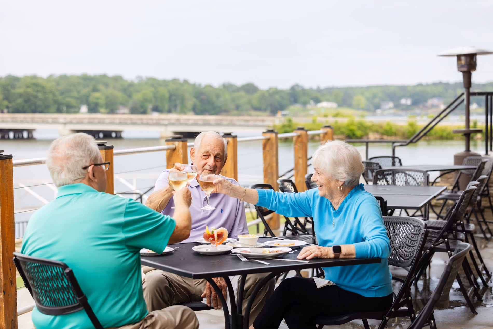 Three older adults sitting outside at a table, toasting drinks together and smiling.