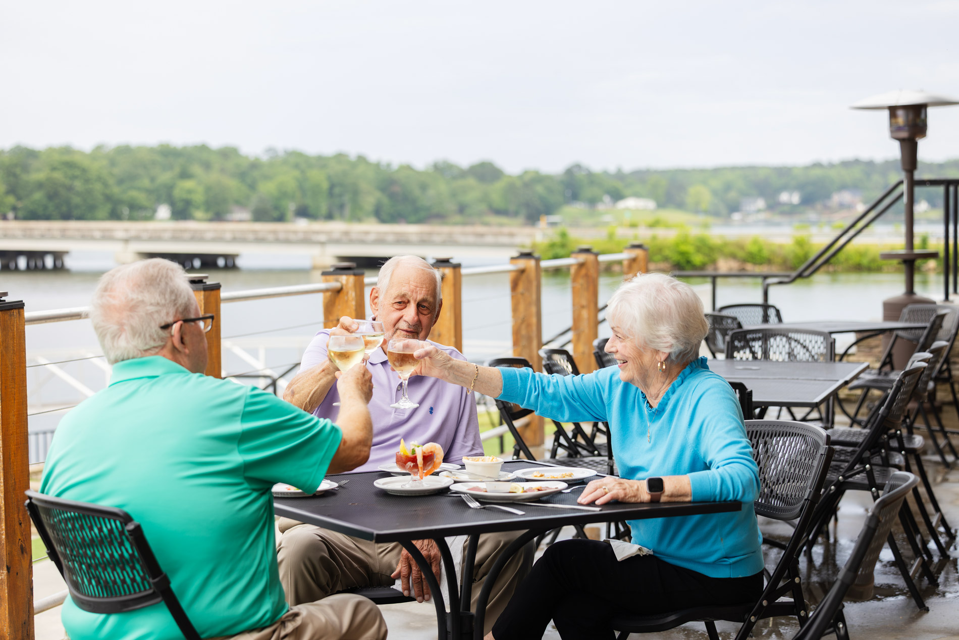 Three older adults sitting outside at a table, toasting drinks together and smiling.