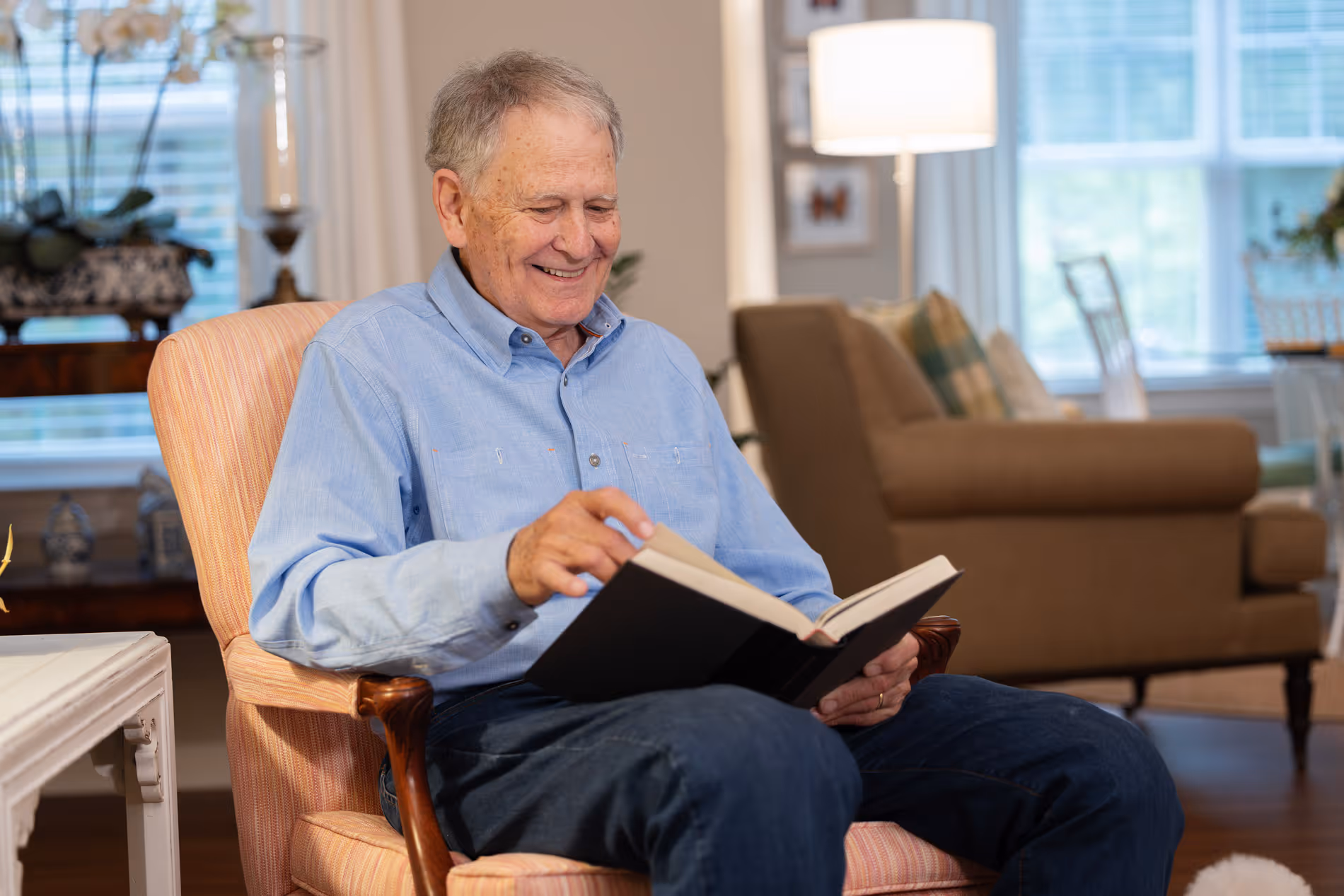 An elderly man sits in an armchair, smiling while reading a book in a cozy living room.