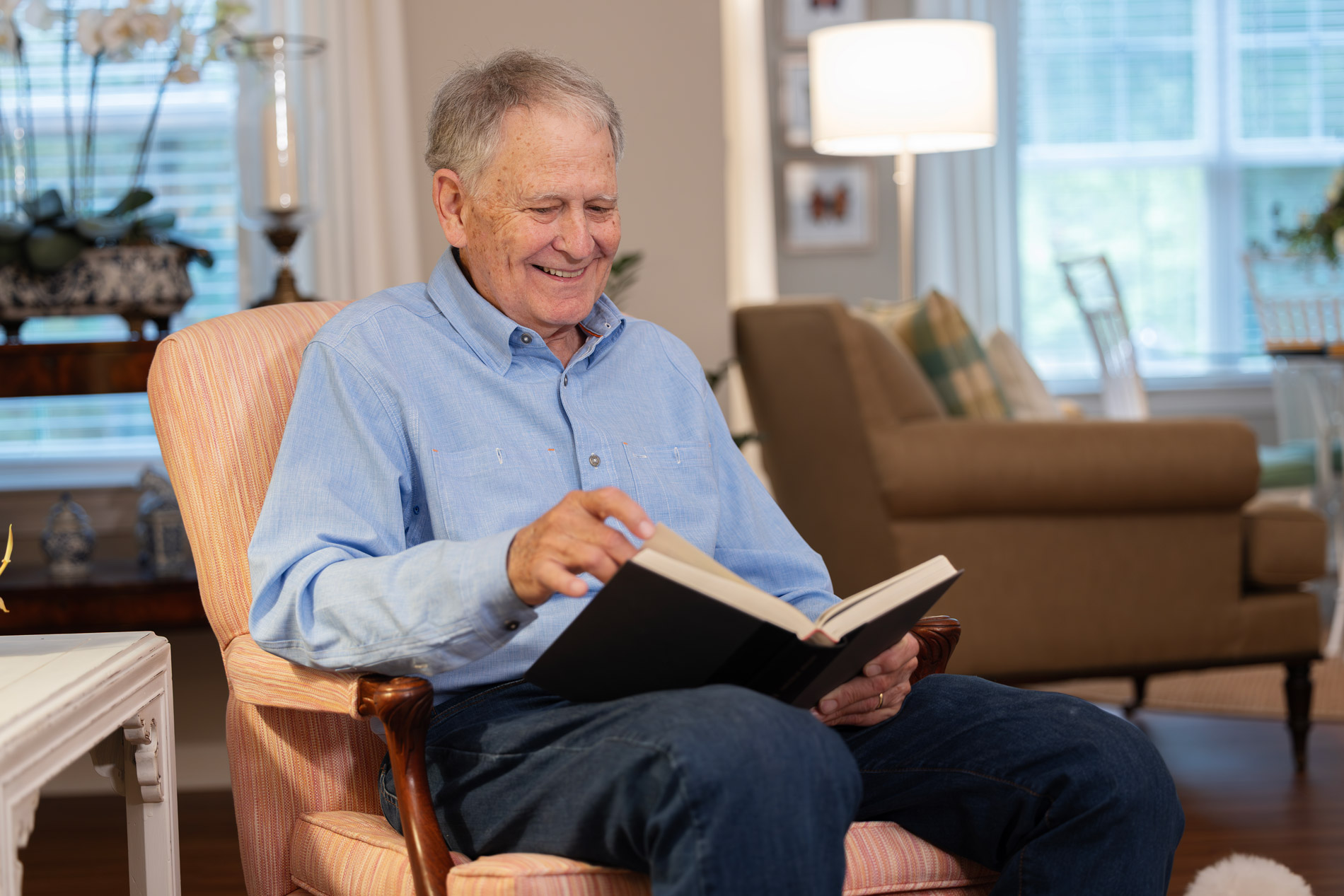 An elderly man sits in an armchair, smiling while reading a book in a cozy living room.