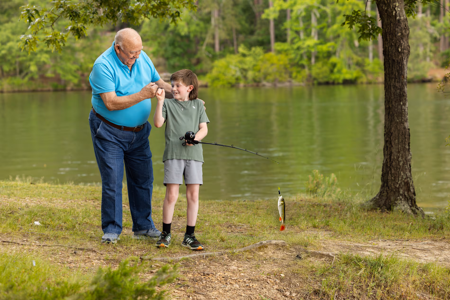 An older man and a young boy celebrate after catching a fish by a lake.