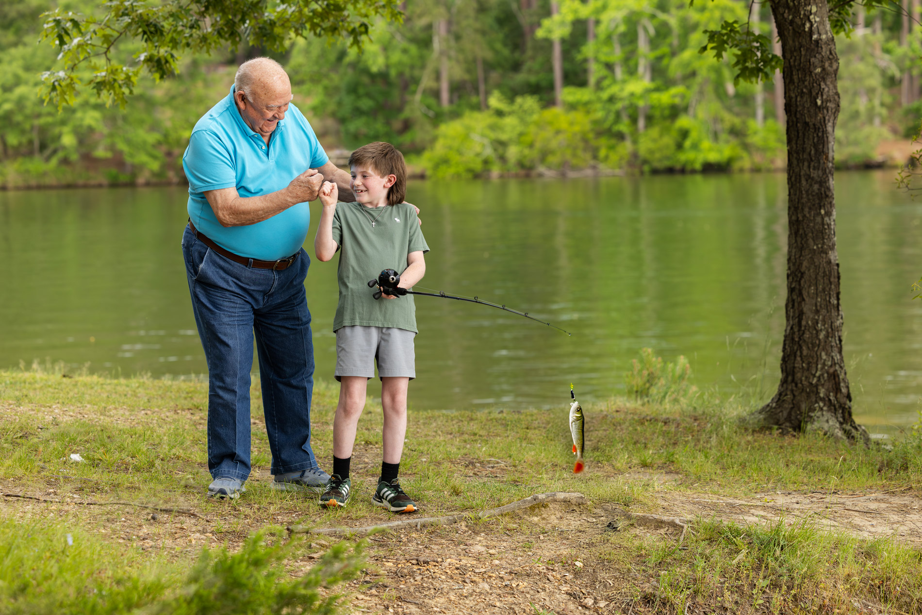 An older man and a young boy celebrate after catching a fish by a lake.