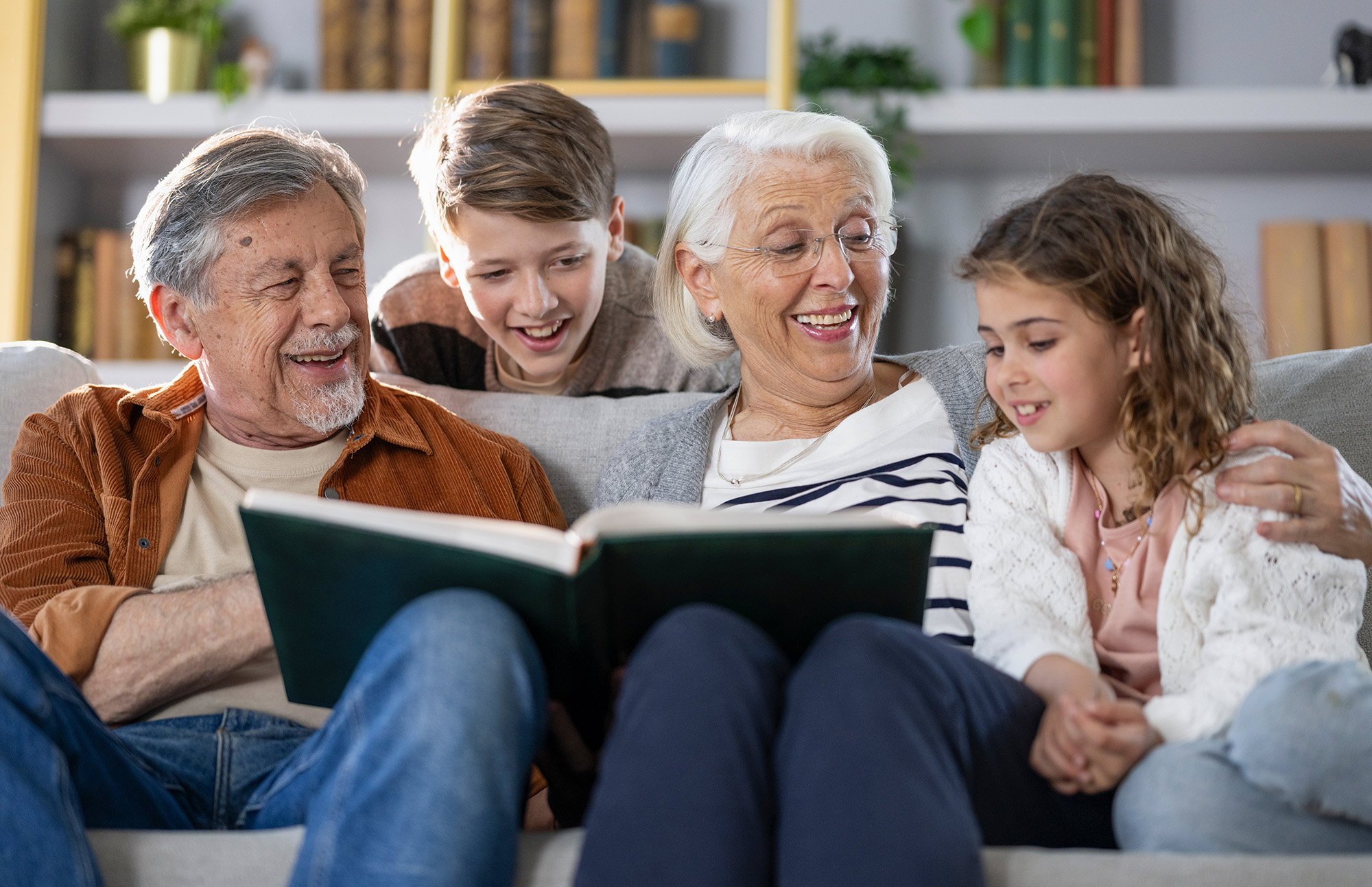 Elderly couple and two children sitting on a couch, happily looking at a book together.