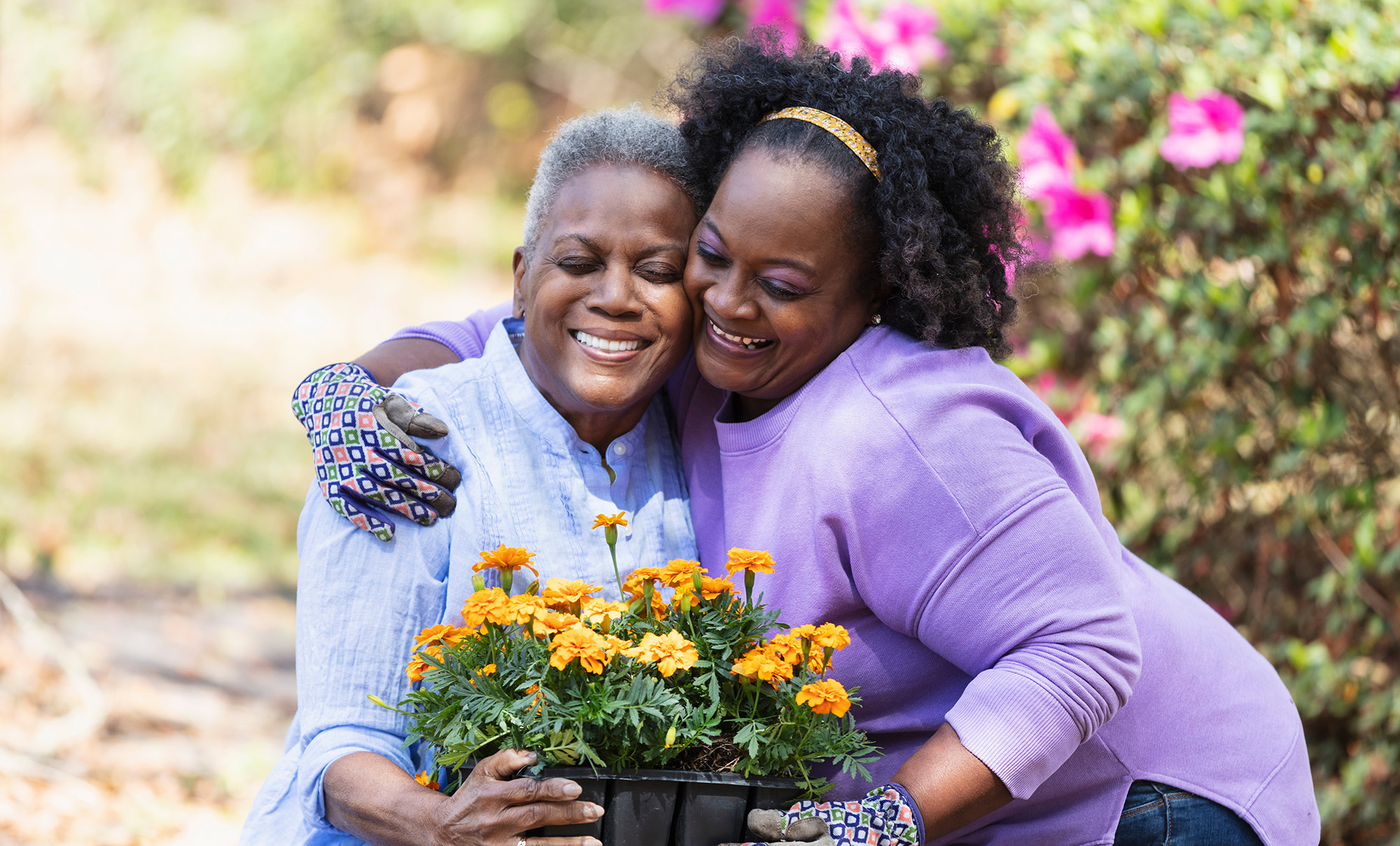 Two women happily hugging while holding a pot of yellow flowers outdoors.