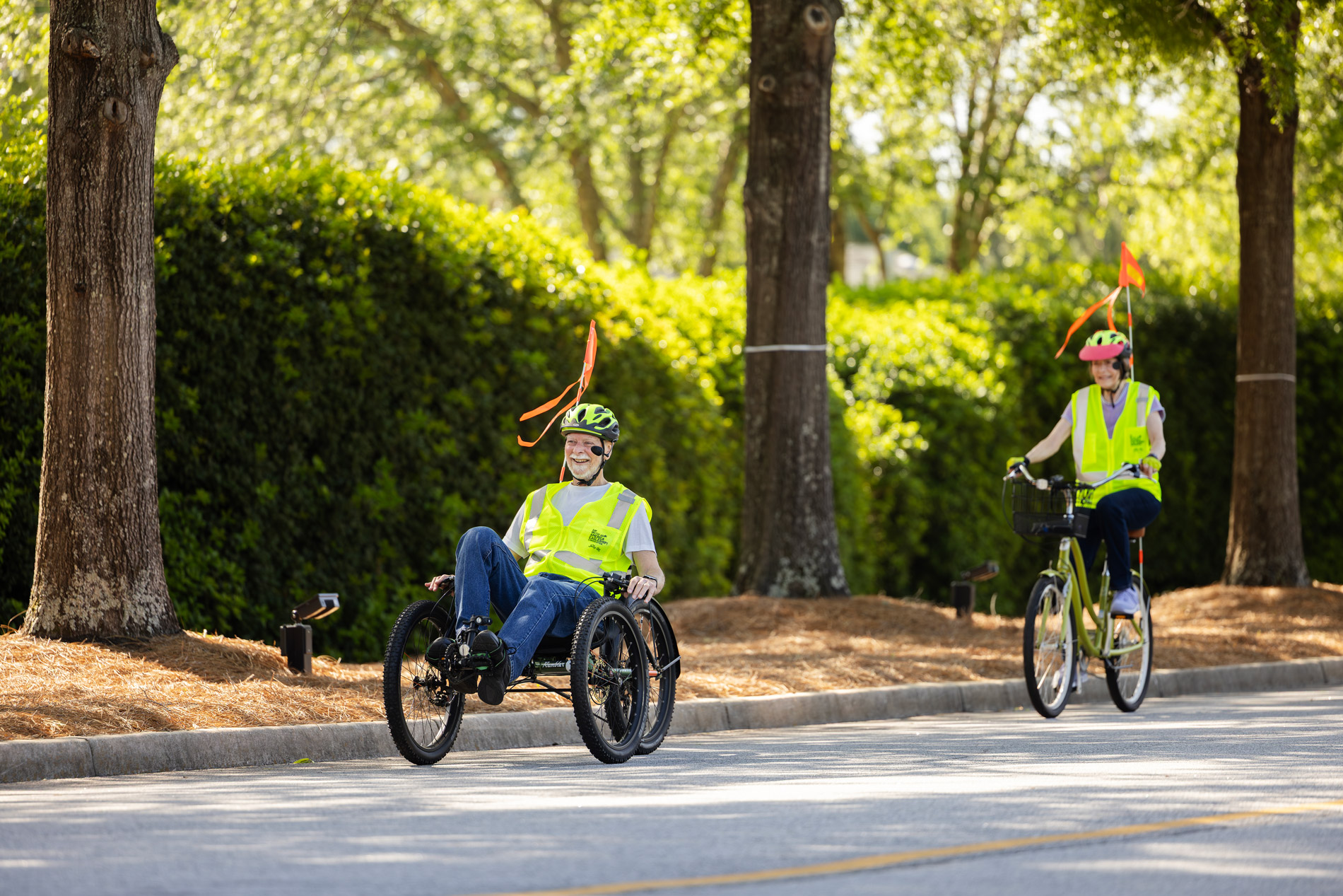 Two people in safety vests ride a tricycle and bicycle on a tree-lined street.