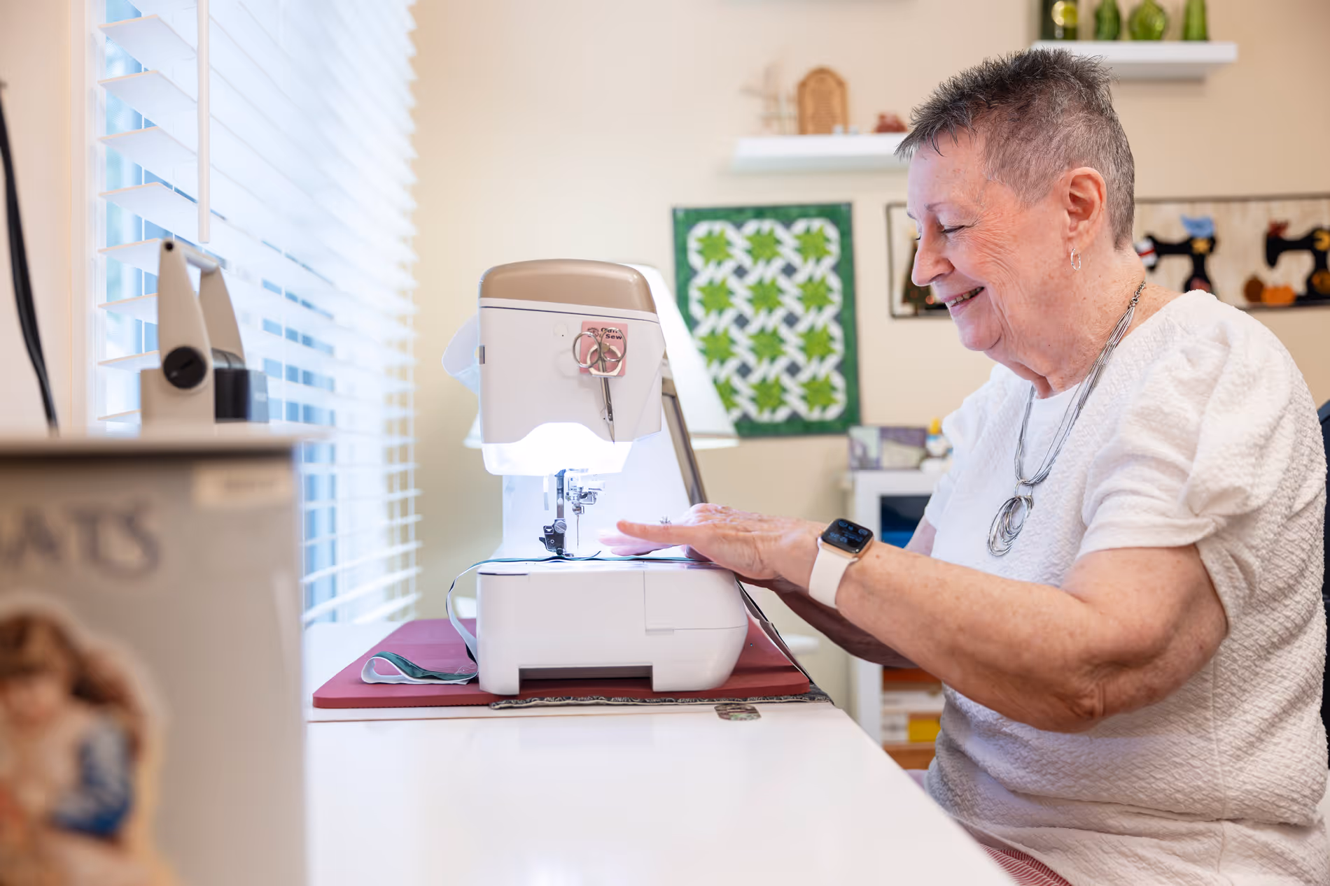 An elderly woman smiles while sewing fabric on a sewing machine near a window.