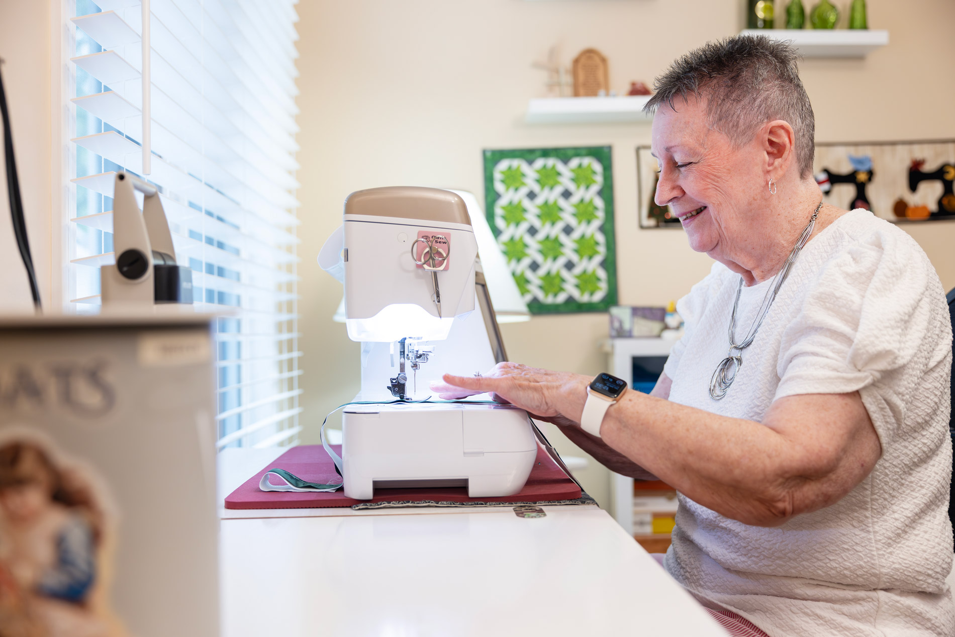 An elderly woman smiles while sewing fabric on a sewing machine near a window.