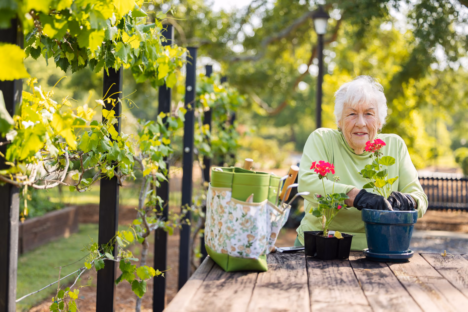An elderly woman smiles while planting flowers at a wooden table in a garden.