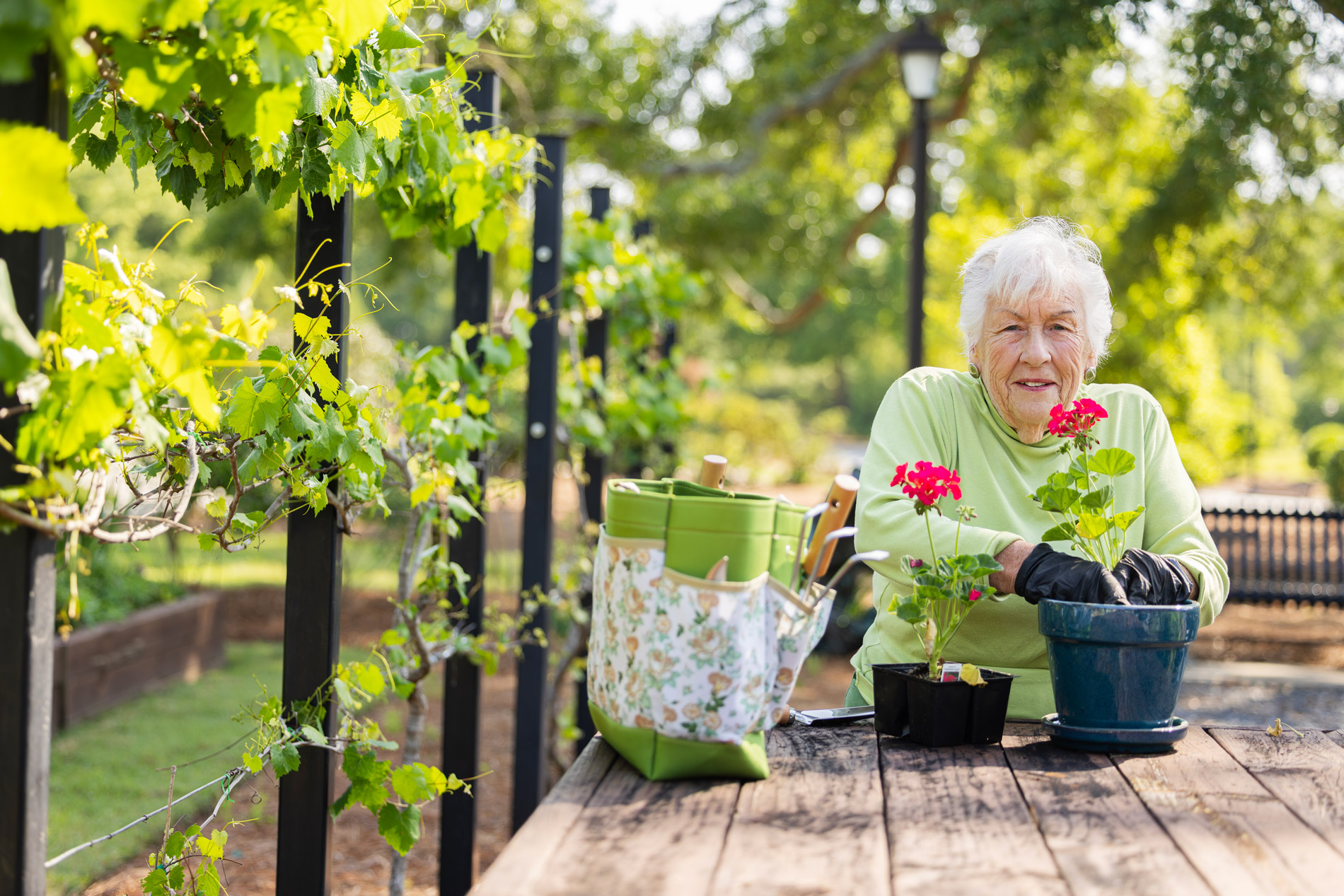 An elderly woman smiles while planting flowers at a wooden table in a garden.