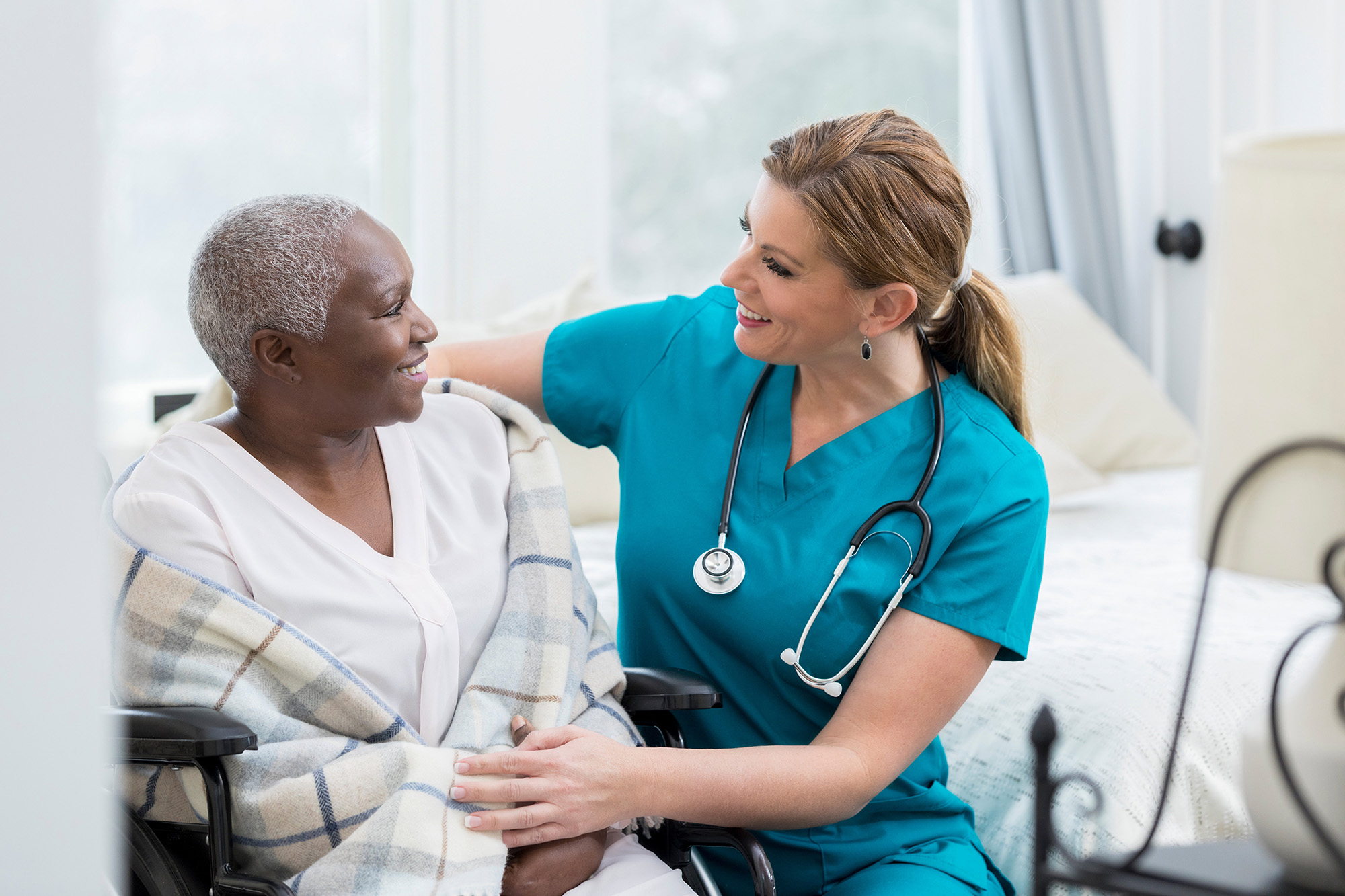 A nurse in scrubs smiles supportively at a seated patient wrapped in a plaid blanket.