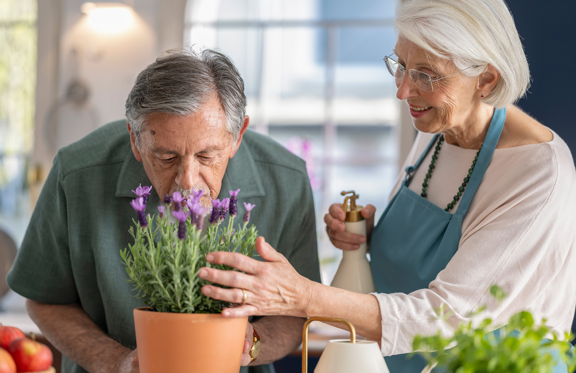 An elderly man smells flowers in a pot while a woman smiles and holds a small watering can.