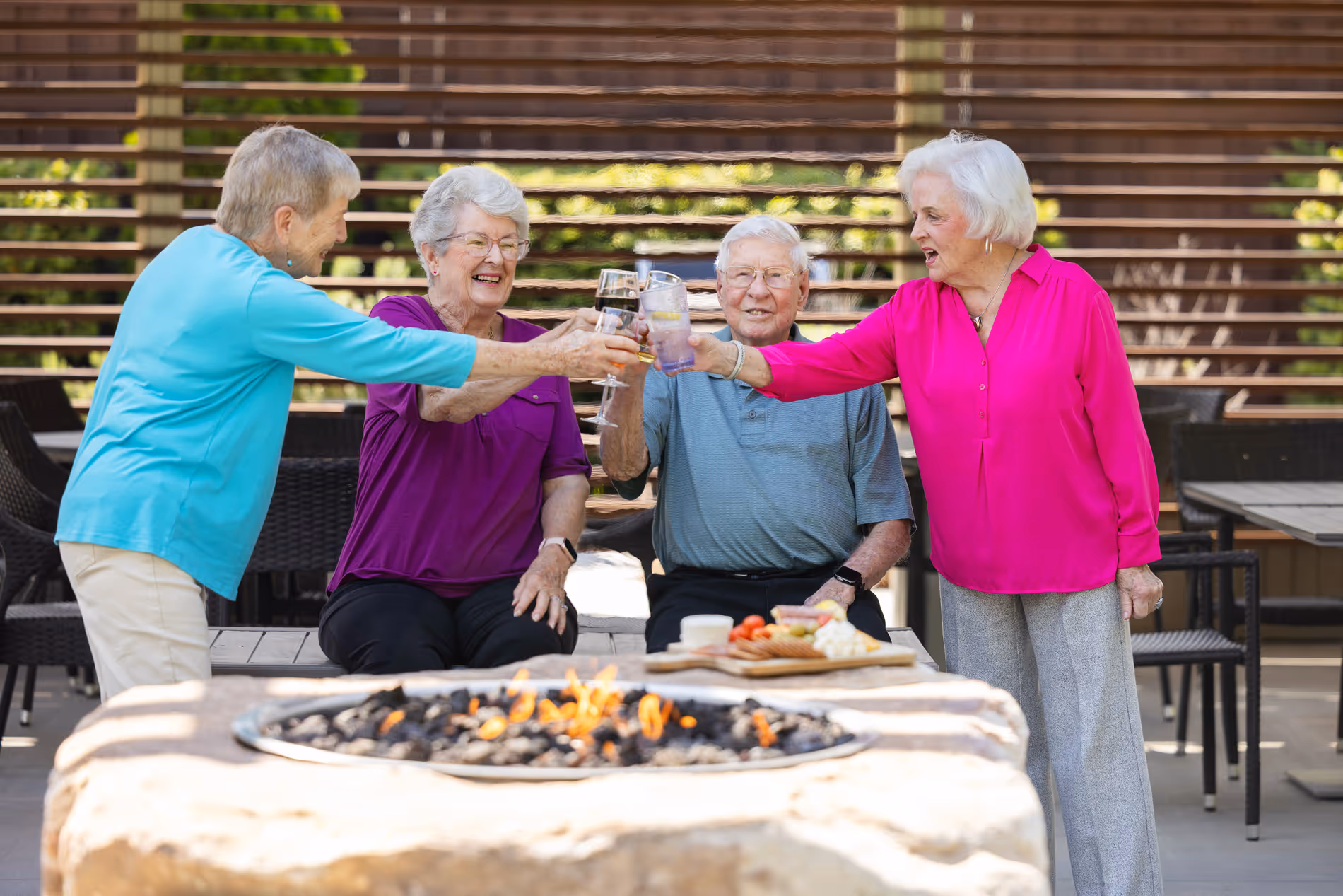 Four elderly people clink glasses together while sitting around a fire pit outdoors.