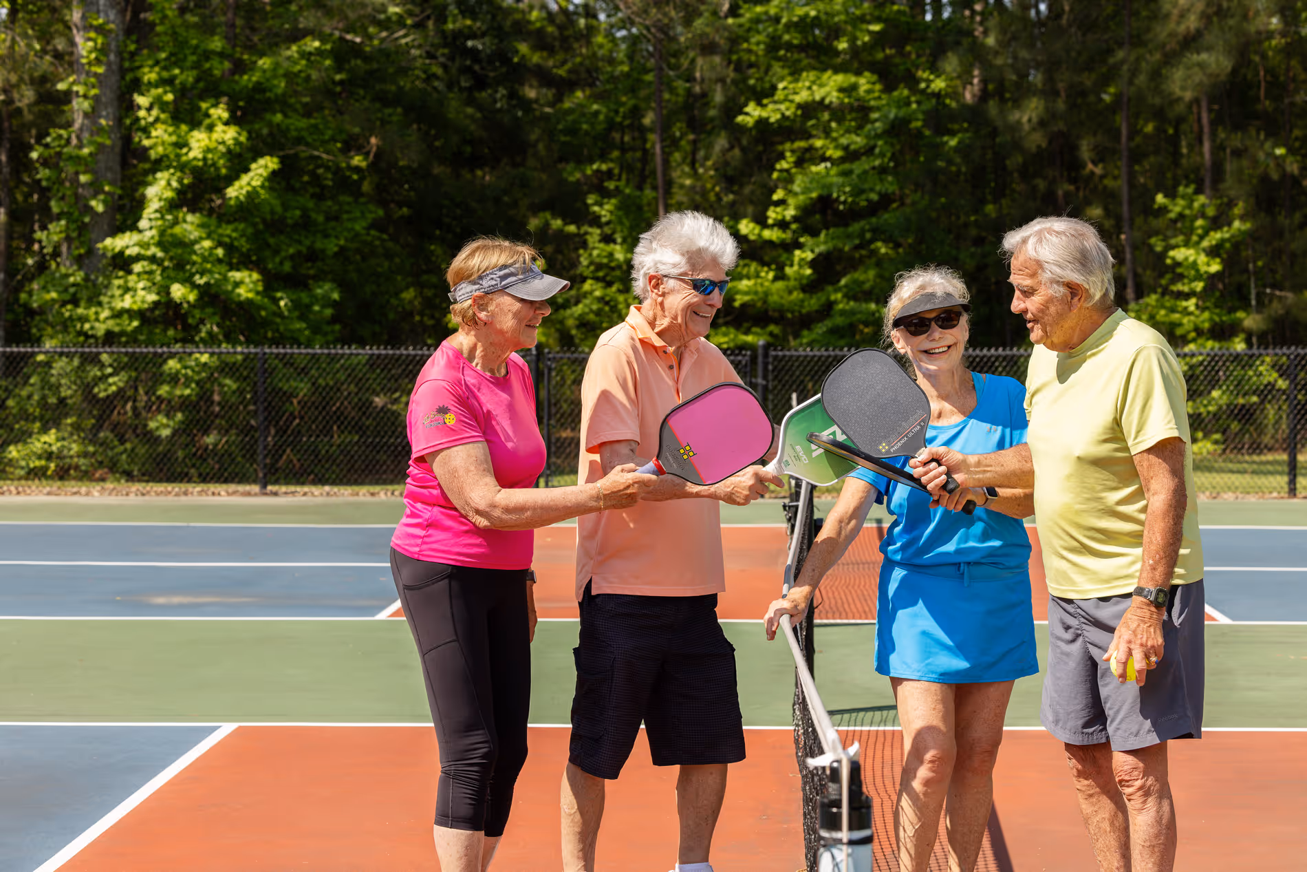 Four senior adults standing on a colorful outdoor pickleball court, smiling and holding paddles together over the net.