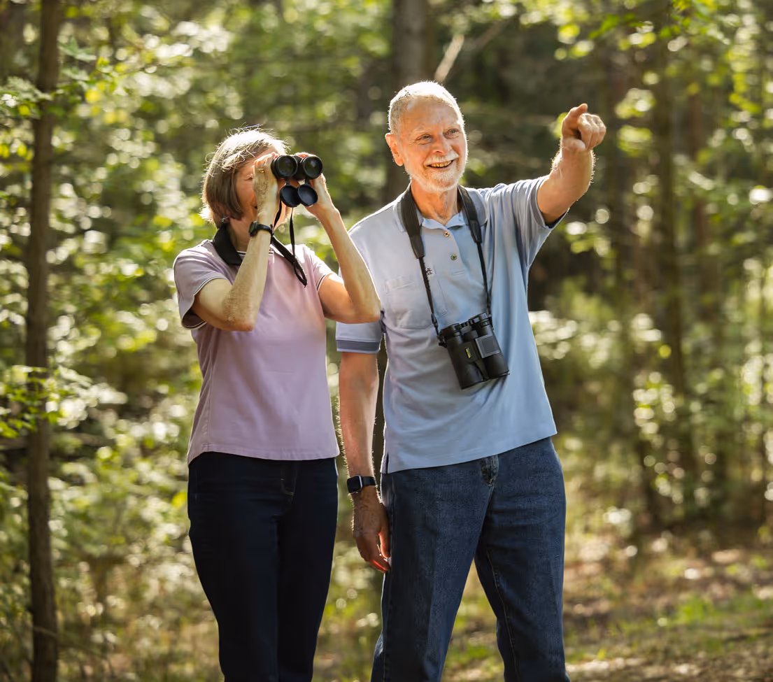 Elderly man and woman birdwatching in a forest, with the woman using binoculars and the man pointing ahead.