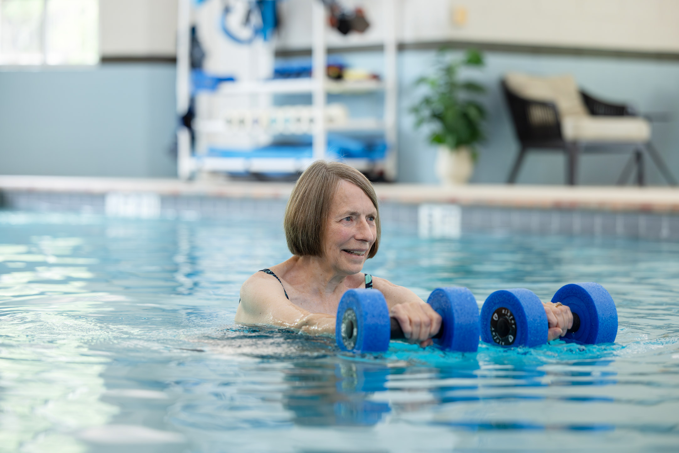 Senior woman exercising in a swimming pool with blue foam dumbbells.