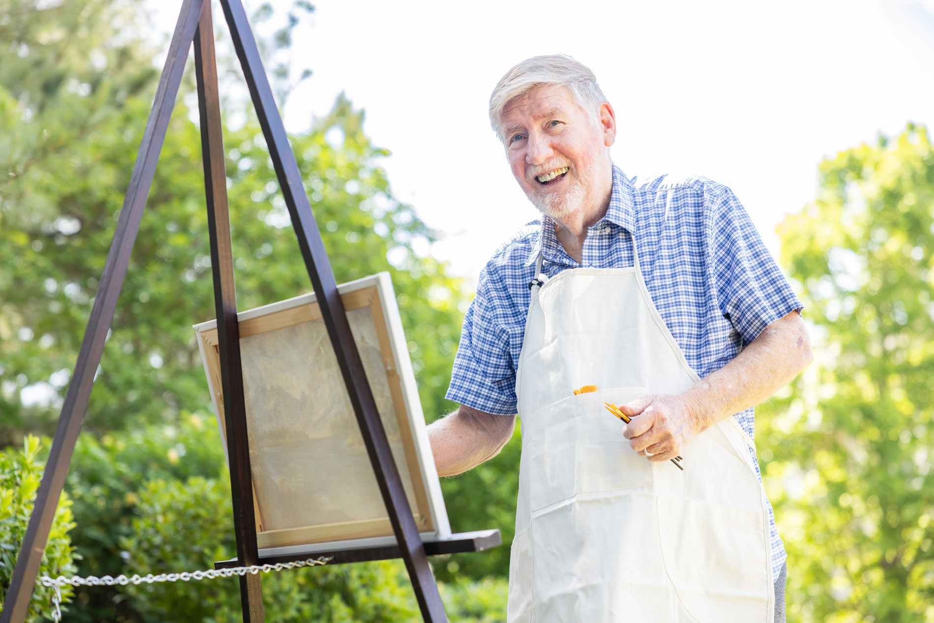 Elderly man outdoors wearing an apron, smiling while painting on an easel.