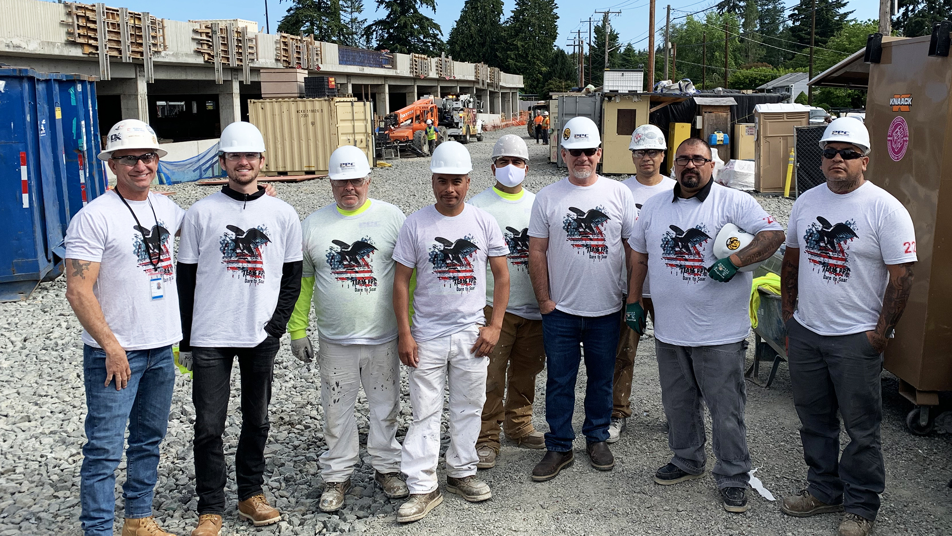 Eight construction workers in matching white shirts, helmets, and gloves stand together on a gravel site, conveying teamwork. Construction materials and equipment are visible in the background, suggesting an active work environment.
