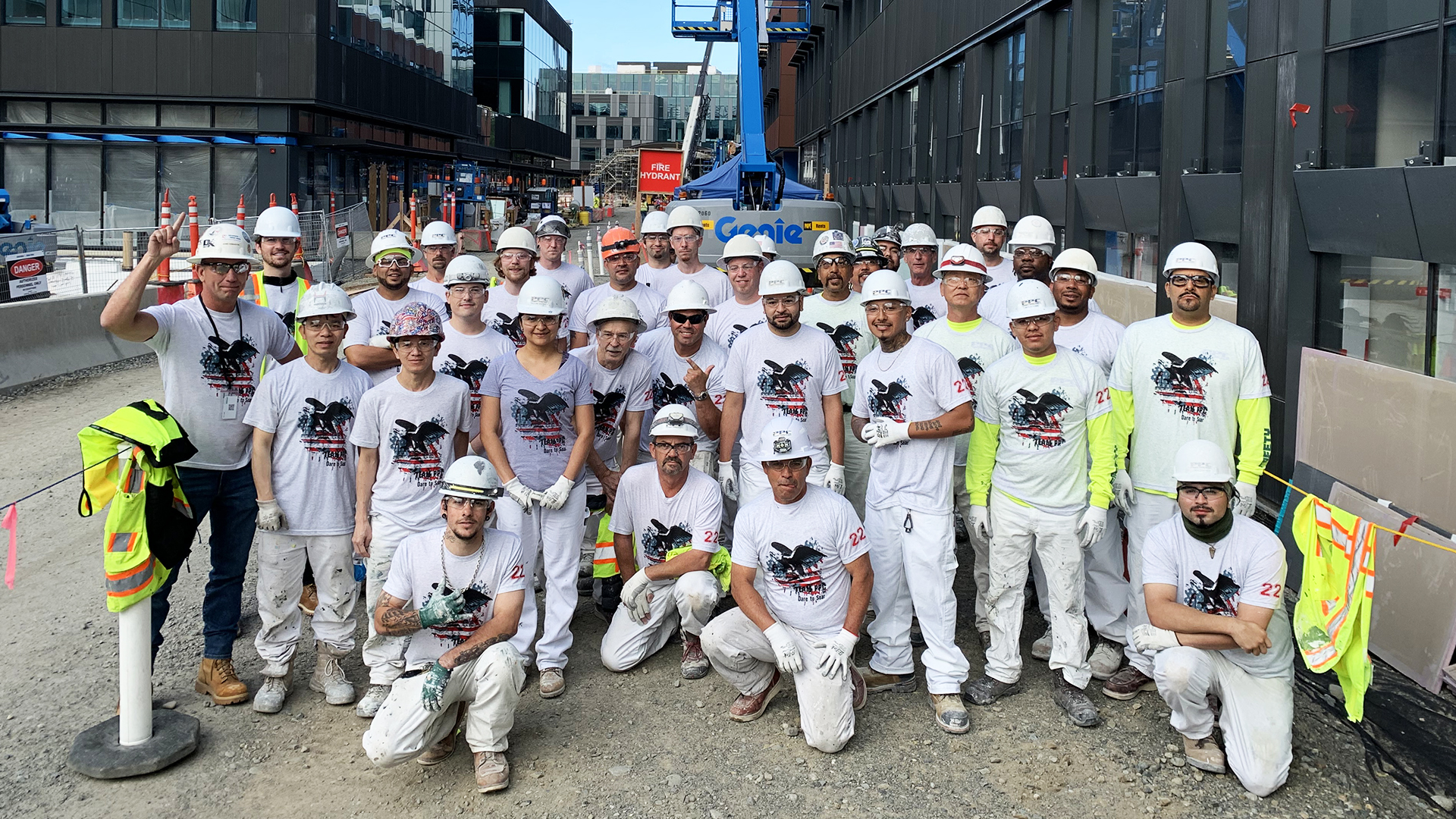 Group of construction workers wearing helmets and matching shirts with a graphic design, posing on a construction site with a positive, unified vibe.