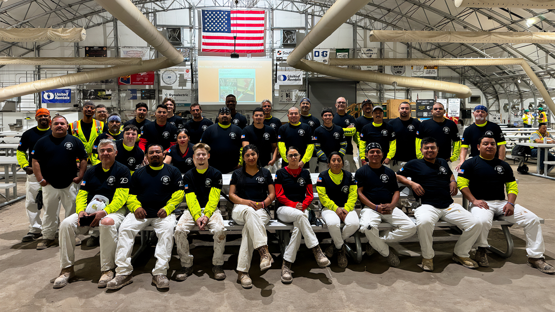 A large group of construction workers in black shirts and yellow sleeves pose inside a warehouse. An American flag is displayed in the background, conveying teamwork.