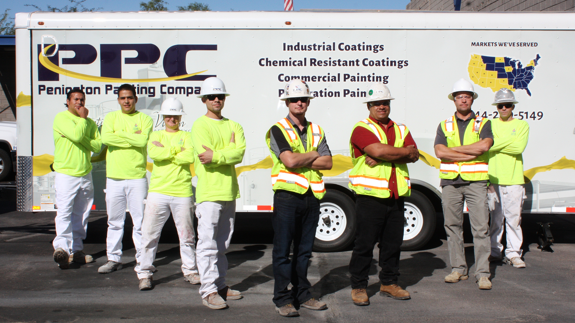 Eight construction workers wearing neon safety gear and helmets stand confidently in front of a company trailer advertising industrial coatings.