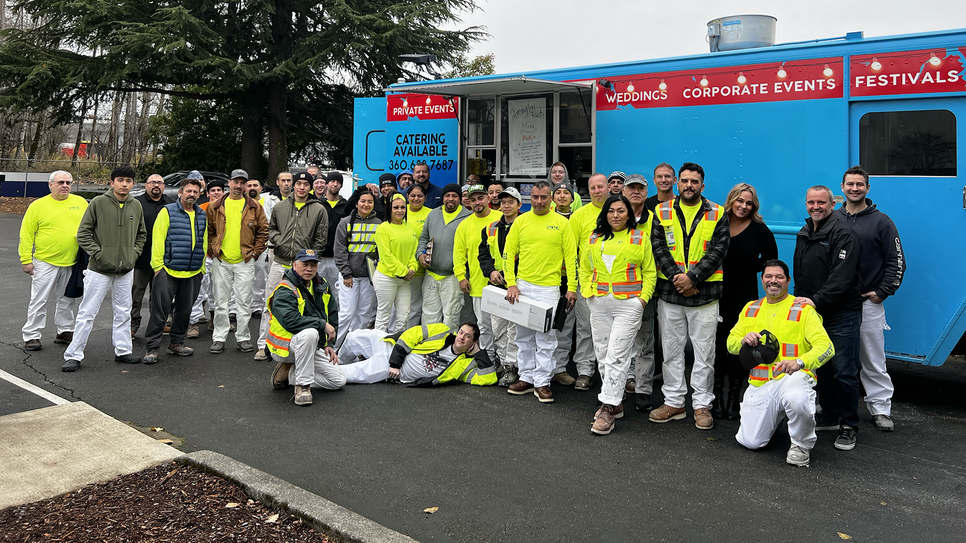 A large group of people, many wearing bright yellow safety vests, stand smiling in front of a blue food truck. The mood is friendly and cheerful.