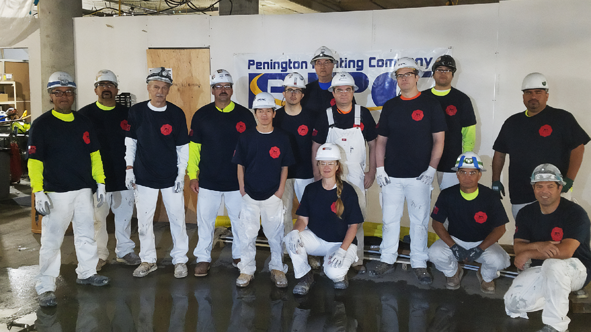 A group of construction workers wearing helmets and matching black shirts with red logos pose together in a building site. The atmosphere is collaborative.