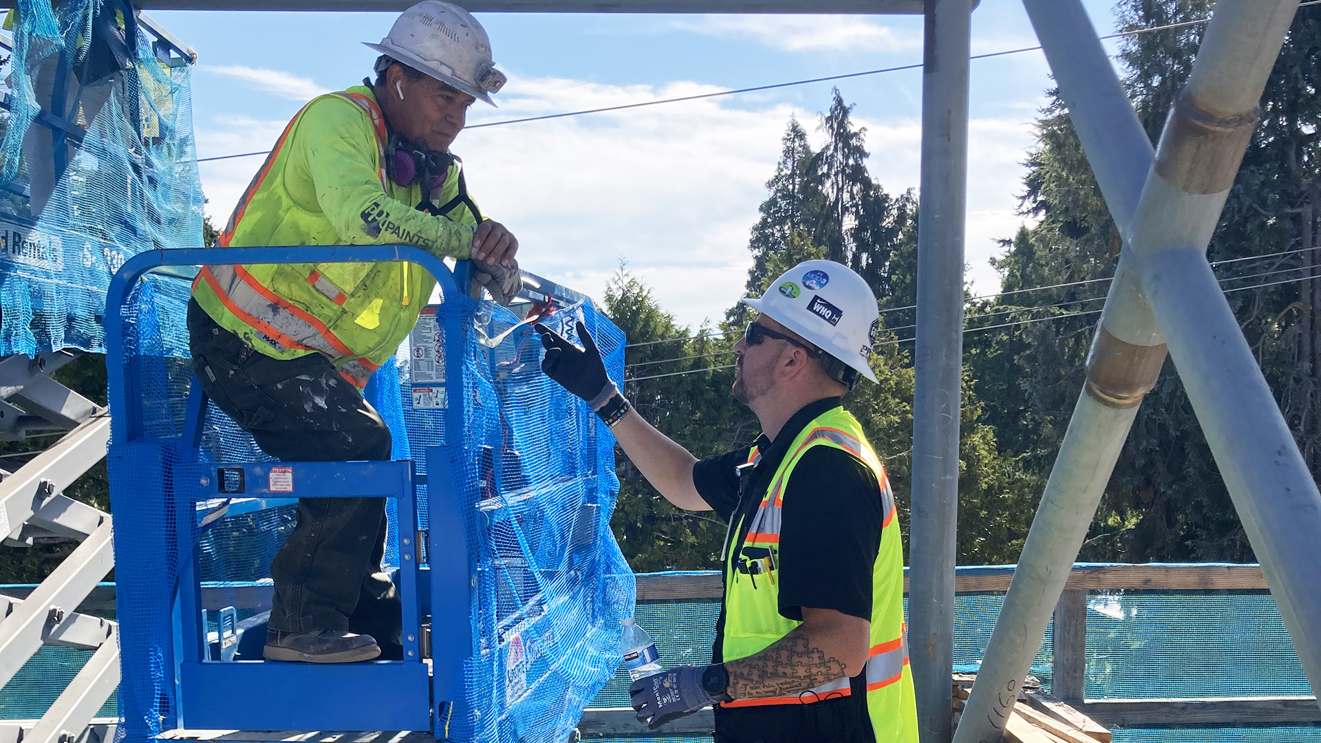 A construction worker in a hard hat and neon vest stands on a blue lift, while another gestures and holds a water bottle. The outdoor scene, with trees and a clear sky, conveys teamwork and communication.