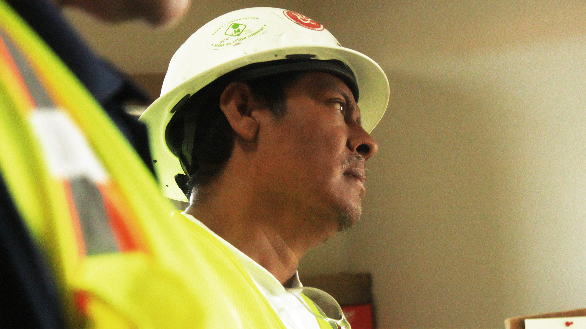 A construction worker wearing a white hard hat and yellow safety vest looks intently forward. The setting suggests a work environment, evoking focus.