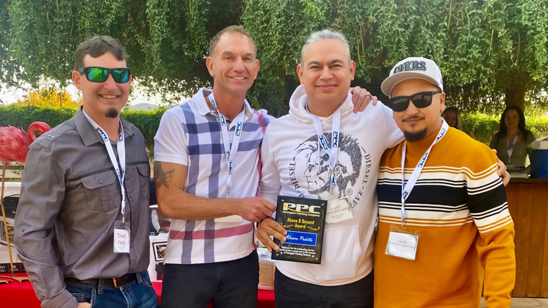 Four men smiling outdoors, one holding an award plaque. They wear casual attire, sunglasses, and name tags. Green foliage and a red flamingo decoration are in the background.