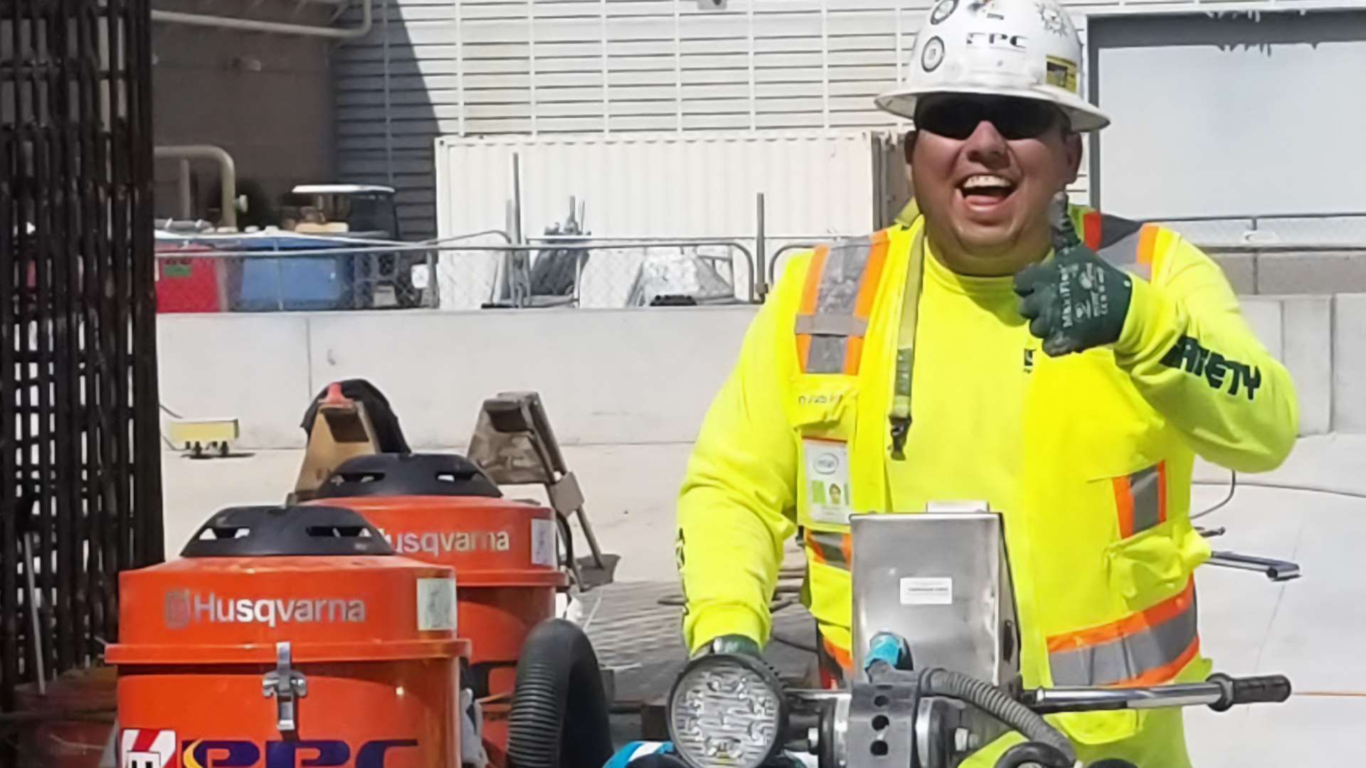 A construction worker in a bright yellow safety vest and hard hat gives a thumbs-up, smiling. The background shows construction equipment and fencing.
