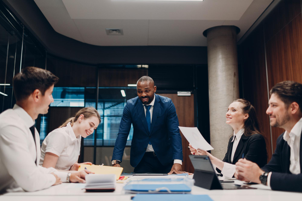 Businessman in blue suit standing and leading a meeting with four colleagues seated at a conference table.
