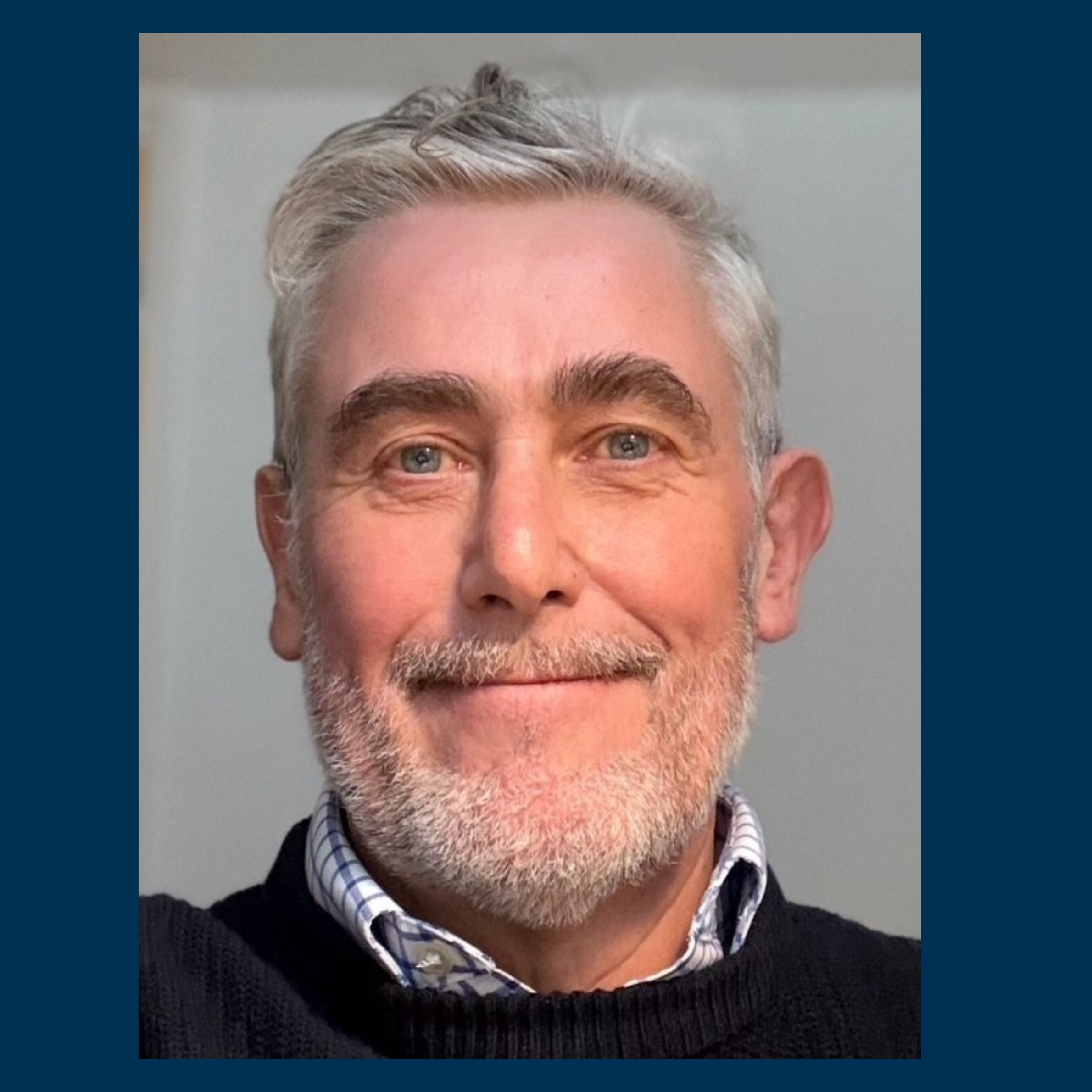 Smiling middle-aged man with gray hair and beard wearing a black sweater over a checkered shirt.