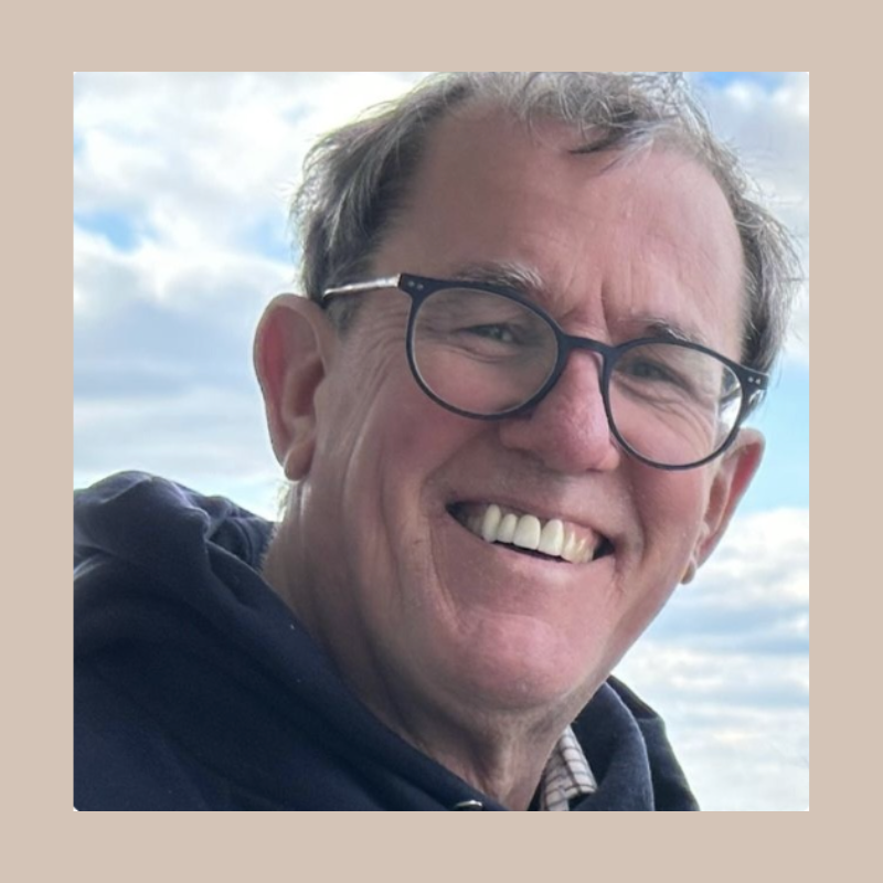 Smiling middle-aged man with glasses wearing a dark hoodie outdoors under a cloudy sky.