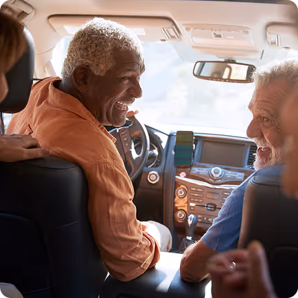 Two elderly men are laughing together inside a car, facing each other from the front seats.