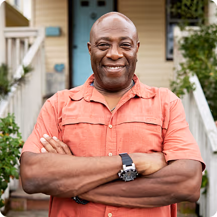 Man in a red shirt stands on a porch, smiling with arms crossed.