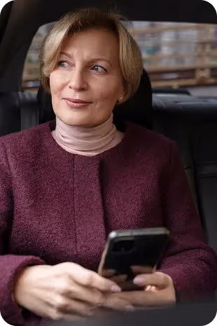 Woman in a purple jacket sitting in a car, holding a smartphone and looking outside.
