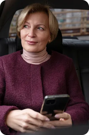 Woman in a purple jacket sitting in a car, holding a smartphone and looking outside.
